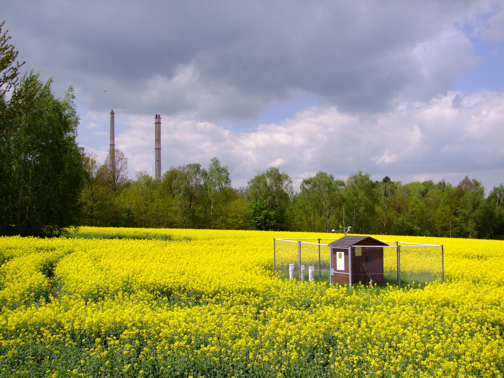 Das Bild zeigt ein eingezäuntes Häuschen mit Messgeräten und zwei Schornsteinen im Hintergrund.