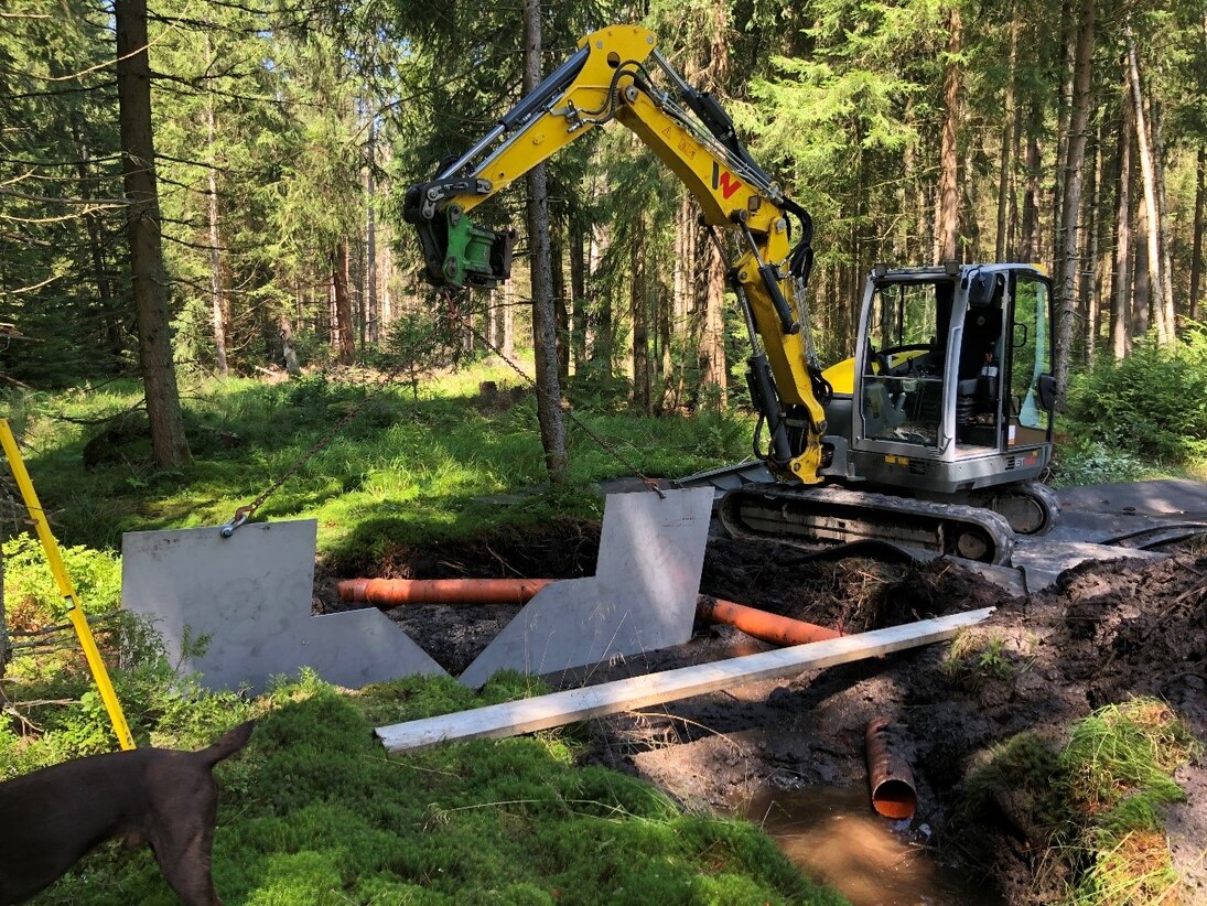 Neben einem Bagger im Wald werden Röhre und V-Wehr in einen Graben installiert.