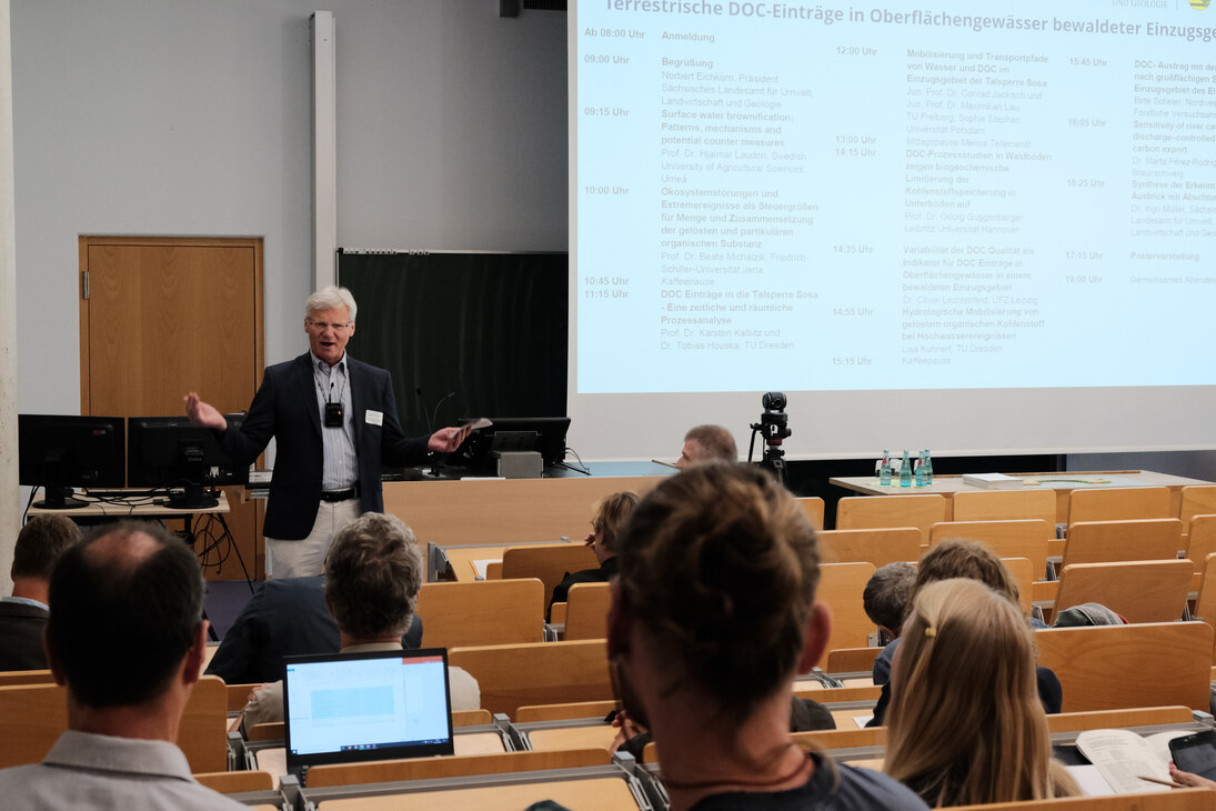 Der Präsident Norbert Eichkorn hält das Begrüßungswort aus der Sicht der Zuschauer. Im Hintergrund befindet sich die Leinwand mit den Programmpunkten des Workshops.