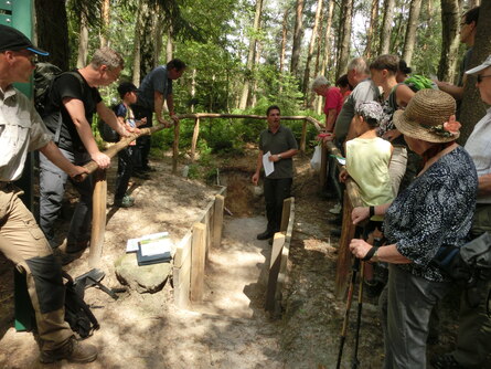 Das Bild zeigt eine Gruppe von Exkursionsteilnehmern um eine mit Holzgeländern abgesperrte Bodengrube im Wald stehend. Ein Mann steht in der Bodengrube und erläutert die Entstehung des Bodens.